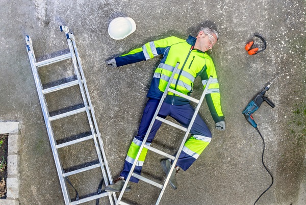 An overhead view of an Ohio construction worker in high-visibility safety gear lying unconscious on the ground next to a fallen silver ladder, a hard hat, and power tools.